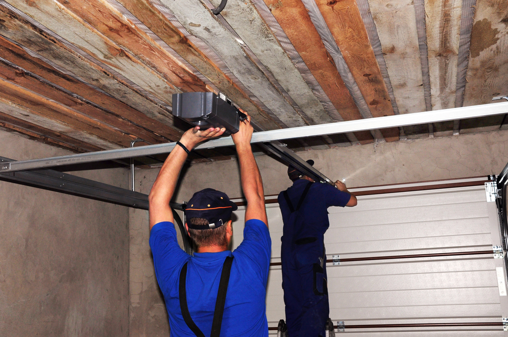 Two workers in blue uniforms install or repair an automatic garage door opener inside a garage with a wooden ceiling.
