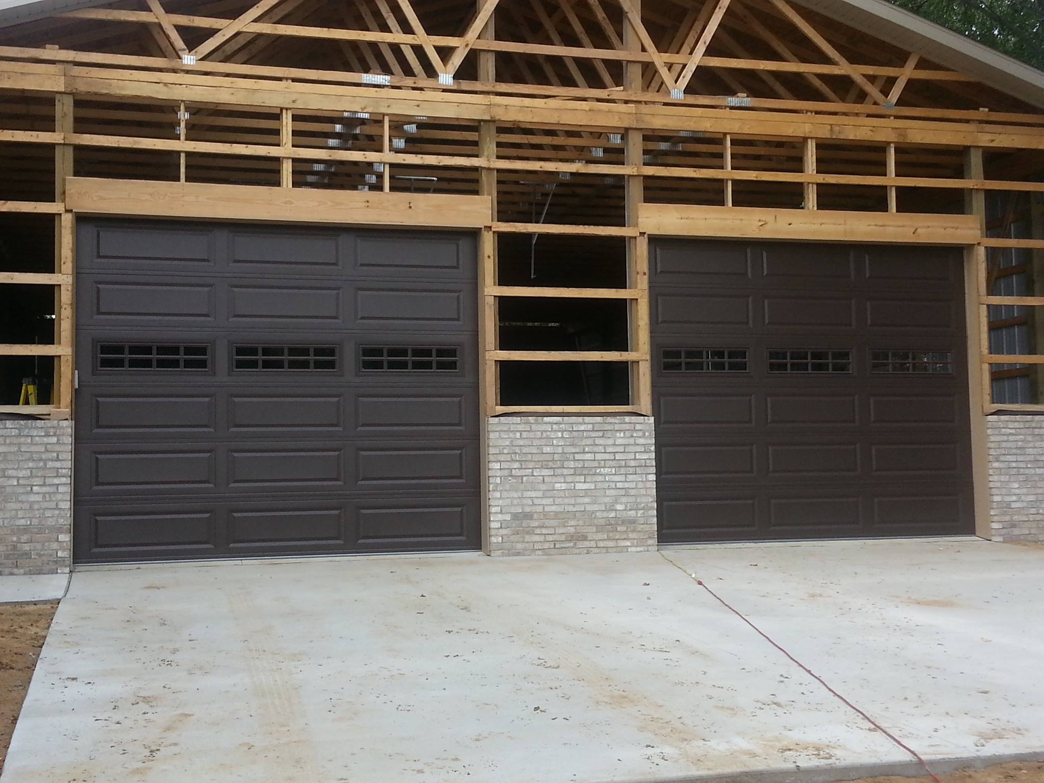 A building under construction with two brown garage doors, exposed wooden framing above, partial brickwork, and a concrete driveway in front.