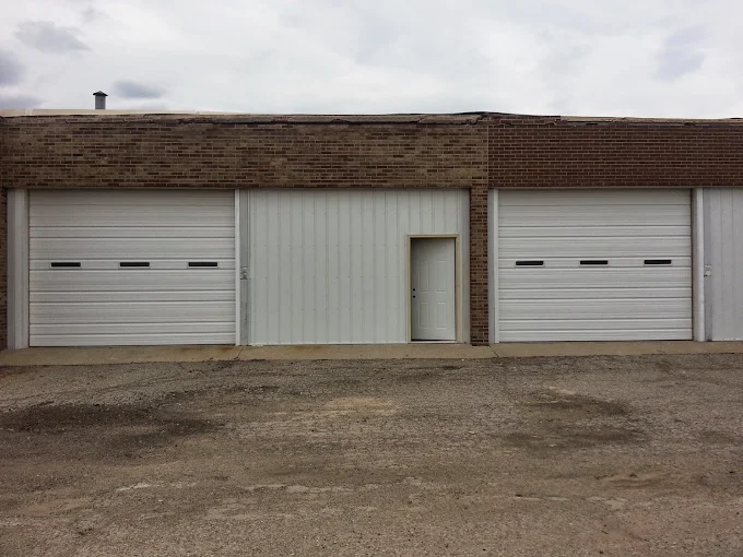 A building with two large white garage doors and a regular-sized door in between them, set in a brick and white metal exterior.