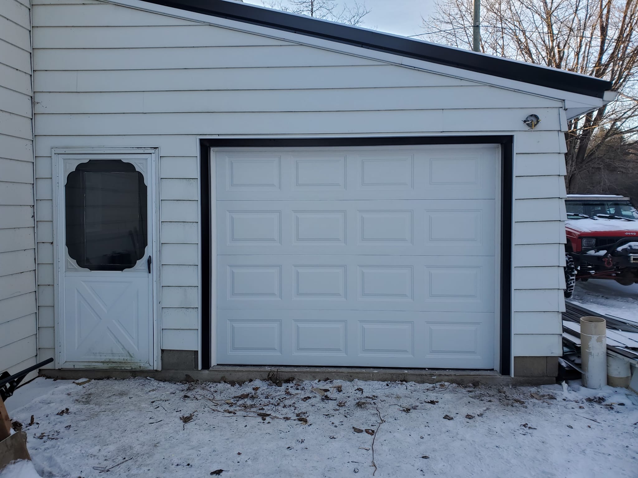 White garage with closed paneled door and adjacent white side door; snow and some debris on the ground; red vehicle partially visible in the background to the right.