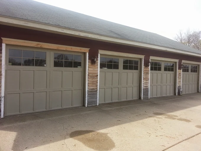 Four closed garage doors with rectangular windows at the top, set in a brown and white building with a concrete driveway in front.