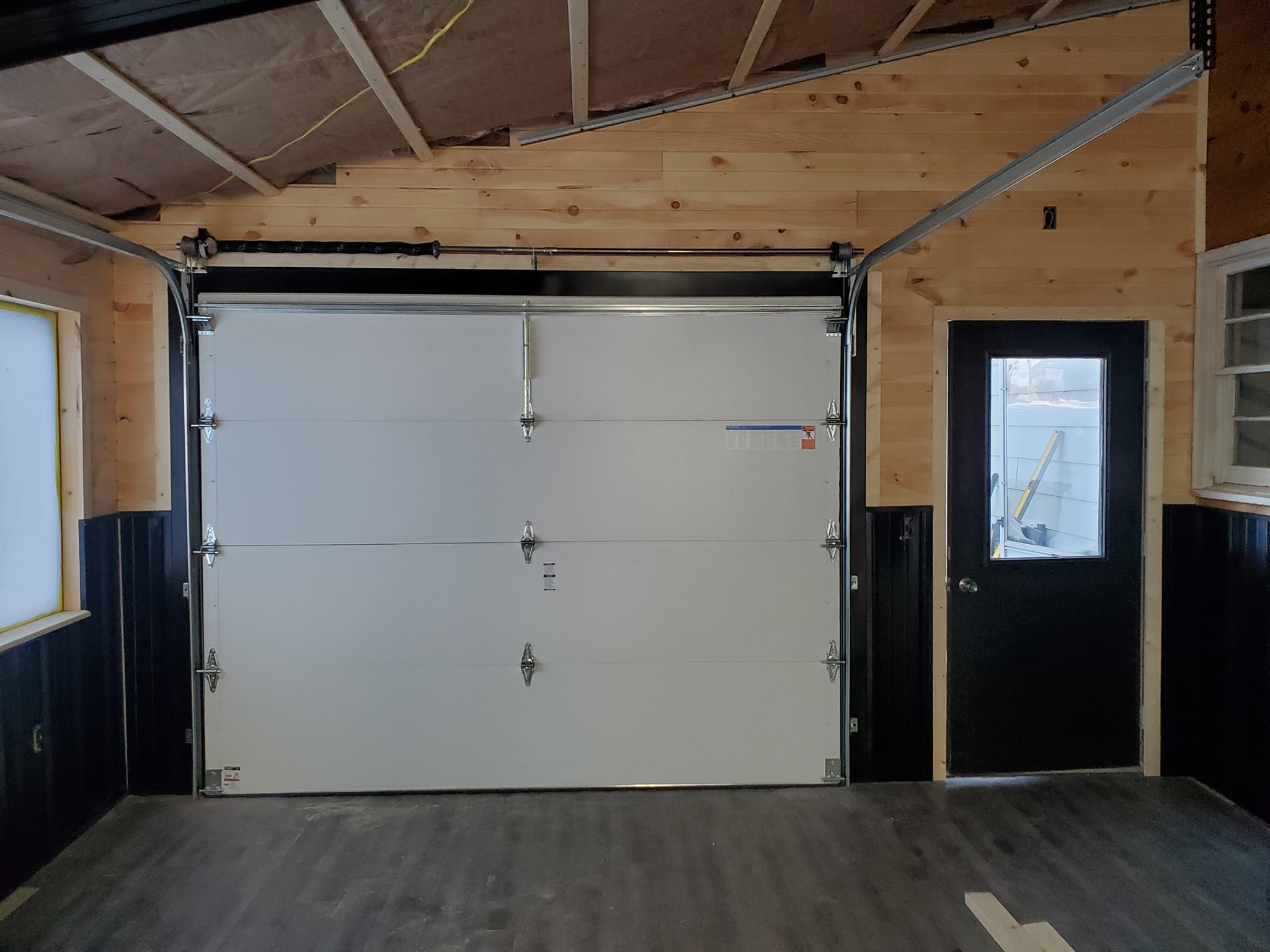 Interior view of a garage with a closed white overhead door, black trim, wood-paneled walls, and a black door with a window to the right.